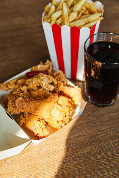 селективный фокус жареной курицы, картошки фри и соды в стекле на деревянном столе при солнечном свете
