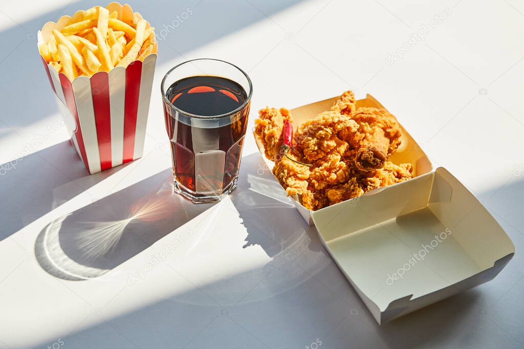 tasty deep fried chicken, french fries and soda in glass on white table in sunlight