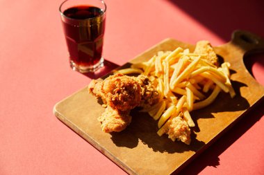 Kızarmış tavuk ve patates kızartması ahşap kesme tahtasında güneş ışığında soda ile servis ediliyor.