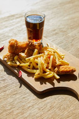 spicy deep fried chicken, french fries on board with soda in glass on wooden table in sunlight clipart