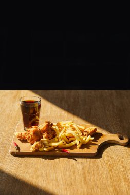 Baharatlı, yağda kızartılmış tavuk, gemide patates kızartması, cam bardakta soda, ahşap masa üzerinde güneş ışığı, siyah üzerine izole edilmiş.