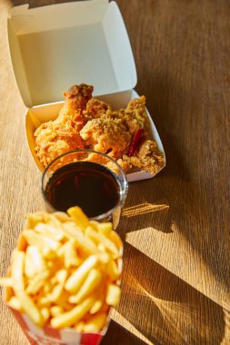 selective focus of spicy deep fried chicken, french fries and soda in glass on wooden table in sunlight clipart