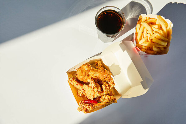 top view of tasty deep fried chicken, french fries and soda in glass on white table in sunlight