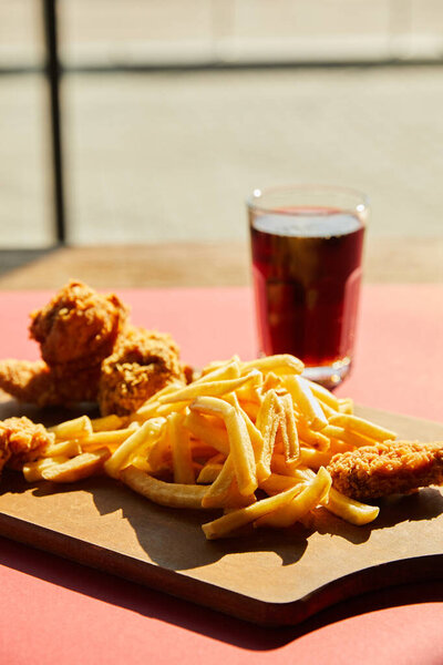 селективный фокус хрустящей жареной курицы и картошки фри подается на деревянной доске с содой в солнечном свете возле окна
