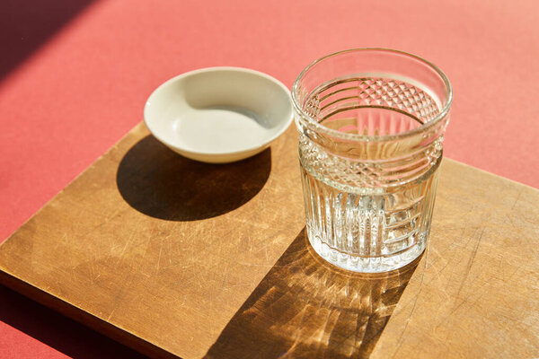 faceted empty glass and white bowl on wooden board in sunlight
