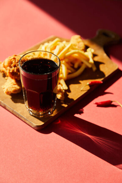 selective focus of spicy deep fried chicken and french fries served on wooden cutting board with soda in sunlight