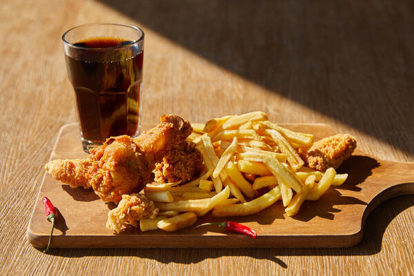 spicy deep fried chicken, french fries on board with soda in glass on wooden table in sunlight