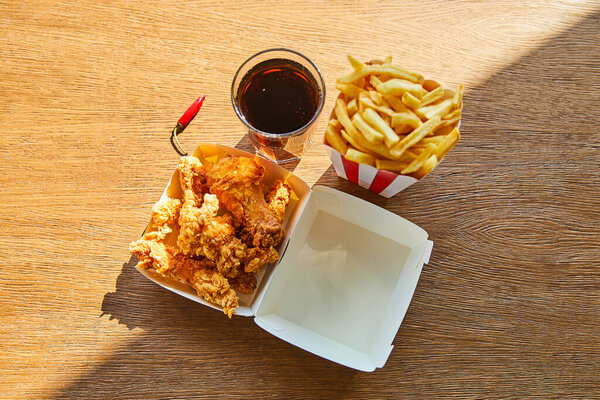 top view of deep fried chicken, french fries and soda in glass on wooden table in sunlight