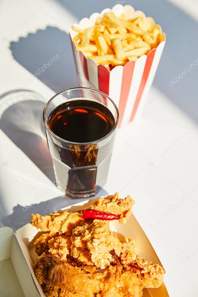 selective focus of tasty deep fried chicken, french fries and soda in glass on white table in sunlight