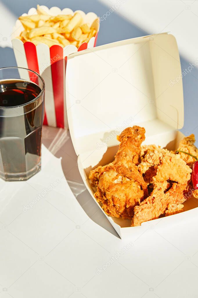 selective focus of tasty deep fried chicken, french fries and soda in glass on white table in sunlight
