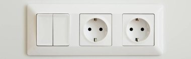 panoramic crop of power sockets near switch on white wall clipart