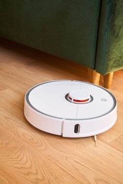 Beyaz robotik süpürge, yeşil kanepenin yanında yerleri yıkıyor.