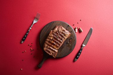 top view of tasty grilled steak served on wooden board on red background with pepper and cutlery clipart