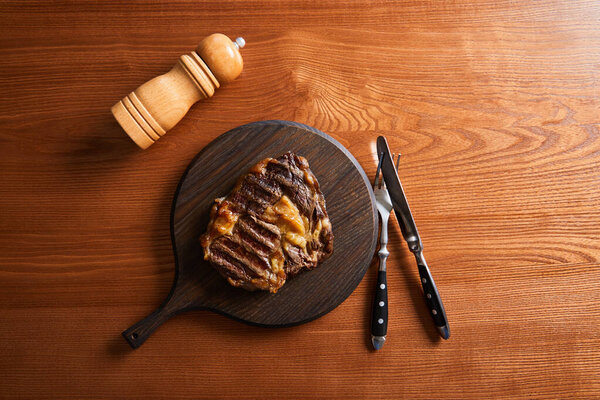 top view of fresh grilled steak on board with cutlery and salt mill on wooden table