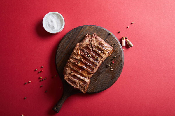 top view of tasty grilled steak served on wooden board on red background with salt and pepper