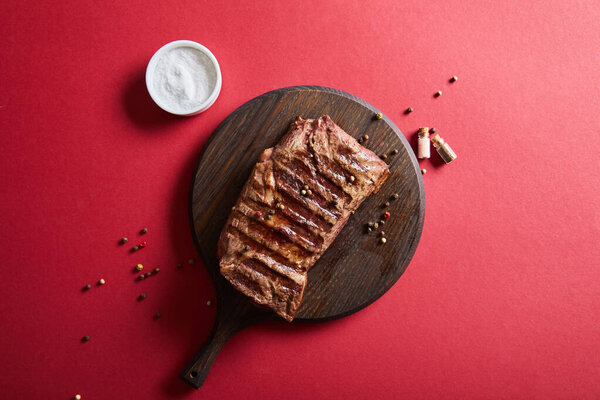 вид сверху на вкусный стейк на гриле подается на деревянной доске на красном фоне с солью и перцем
