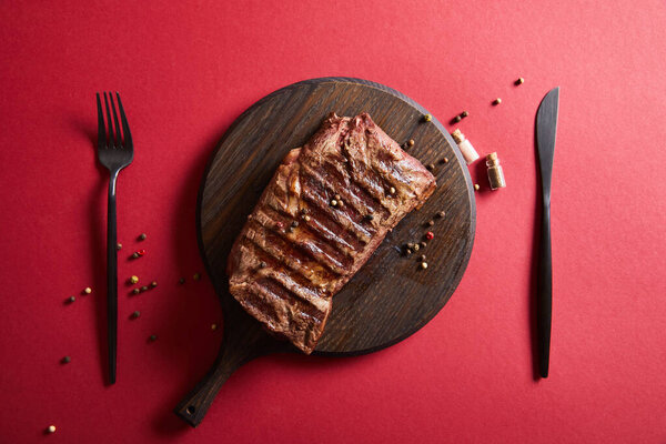 top view of tasty grilled steak served on wooden board on red background with pepper and cutlery