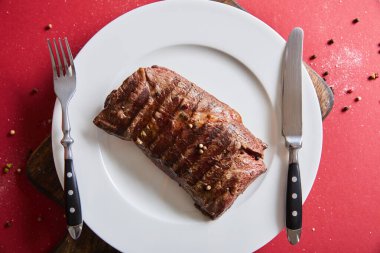Ahşap tabakta servis edilen lezzetli ızgara bifteğin üst görüntüsü kırmızı arka planda biber ve tuzlu.