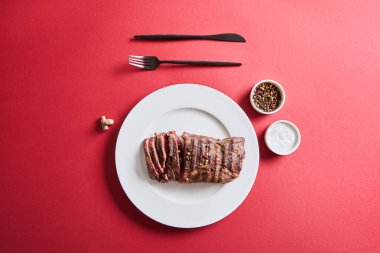 top view of tasty grilled steak served on plate with cutlery and salt and pepper in bowls on red background clipart