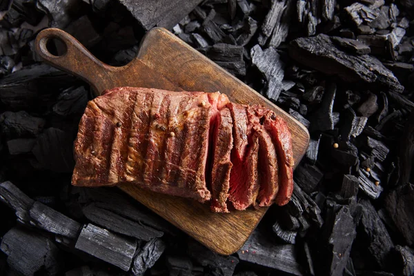 top view of cut fresh grilled tasty steak with rare roasting on wooden cutting board on black coals