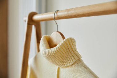 Bej örülmüş yumuşak süveter ahşap askıda asılı, beyazın üstünde.