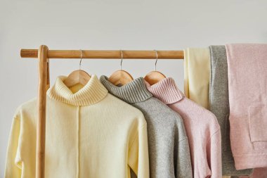 close up view of pink, beige and grey knitted soft sweaters and pants hanging on wooden rack isolated on white clipart