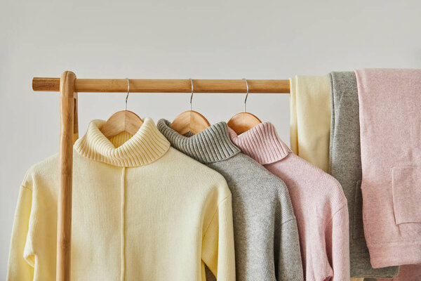 close up view of pink, beige and grey knitted soft sweaters and pants hanging on wooden rack isolated on white