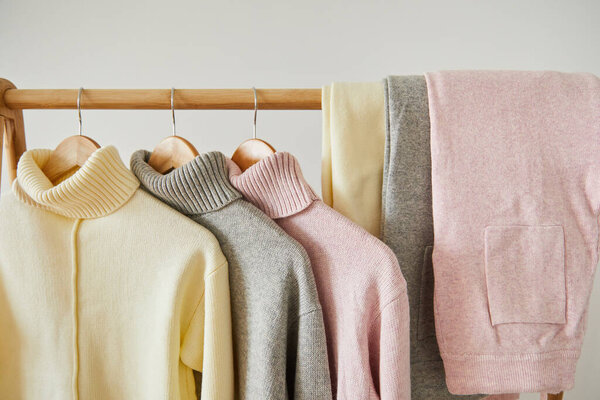 close up view of pink, beige and grey knitted soft sweaters and pants hanging on wooden rack isolated on white