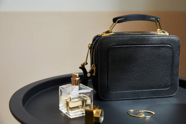 Кожаная сумочка рядом с золотыми серьгами, духи на черном столе на бежевом фоне
