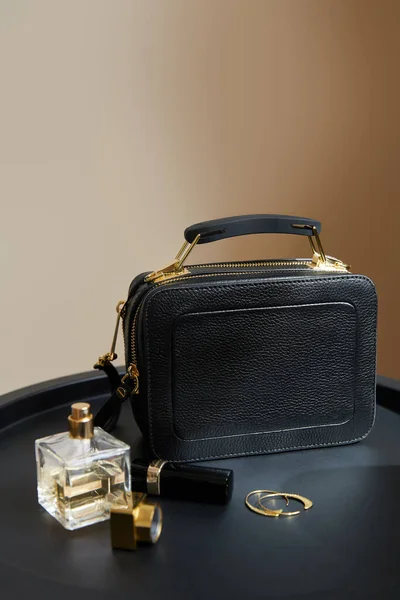 ベージュ地の黒いテーブルの上の黄金のイヤリング 香水や口紅の近くの革ハンドバッグ — ストック写真