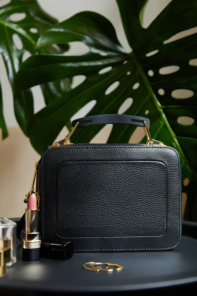 Кожаная сумка рядом с золотыми серьгами и помадой на черном столе возле тропических листьев
