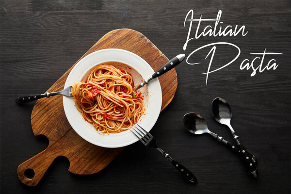 top view of tasty bolognese pasta with tomato sauce in white plate on wooden cutting board near cutlery on black wooden background, italian pasta illustration