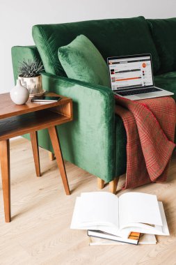 KYIV, UKRAINE - APRIL 14, 2020: green sofa with blanket and laptop with soundcloud website near wooden coffee table with plant and smartphone near books on floor clipart