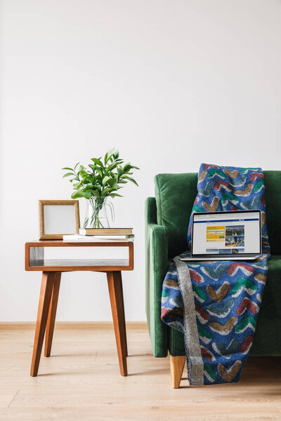 KYIV, UKRAINE - APRIL 14, 2020: green sofa with blanket and laptop with booking website near wooden coffee table with green plant, books and photo frame