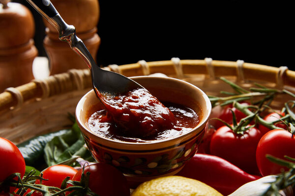 close up view of delicious tomato sauce with spoon near fresh ripe vegetables in basket isolated on black