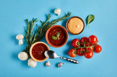 top view of delicious sauces in bowls with spoon near fresh ripe vegetables on blue background clipart