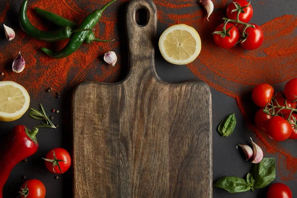 top view of cherry tomatoes, garlic cloves, lemons, green chili peppers, paprika powder and basil leaves near wooden cutting board on black