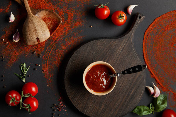 top view of tomato paste in bowl on chopping board near cheery tomatoes and herbs on black