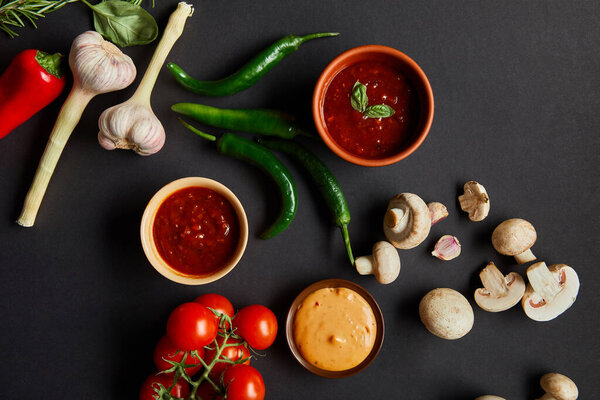 top view of bowls with red tomato and mustard sauces near ripe vegetables on black