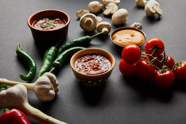 Selective focus of tasty sauces in bowls near ripe cherry tomatoes, green chili peppers, mushrooms and garlic on black — Stock Image Selective Focus Tasty Sauces Bowls Ripe Cherry Tomatoes Green Chili Stock Photo