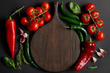 top view of wooden chopping board near ripe cherry tomatoes, garlic cloves, rosemary, basil leaves and green chili peppers on black clipart
