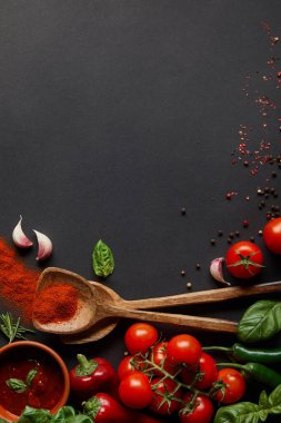 top view of red cherry tomatoes, spicy chili peppers, tomato paste, garlic cloves and fresh herbs near spoons with paprika powder on black  clipart
