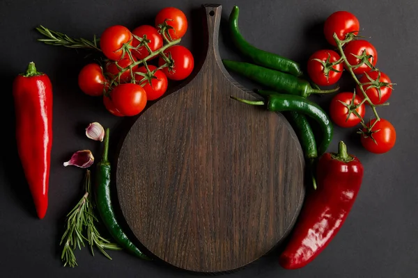 top view of wooden cutting board near ripe cherry tomatoes, garlic cloves, rosemary and green chili peppers on black