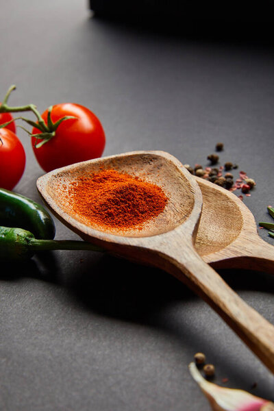 selective focus of organic cherry tomatoes, peppercorns, wooden spoons with paprika powder and green chili peppers on black