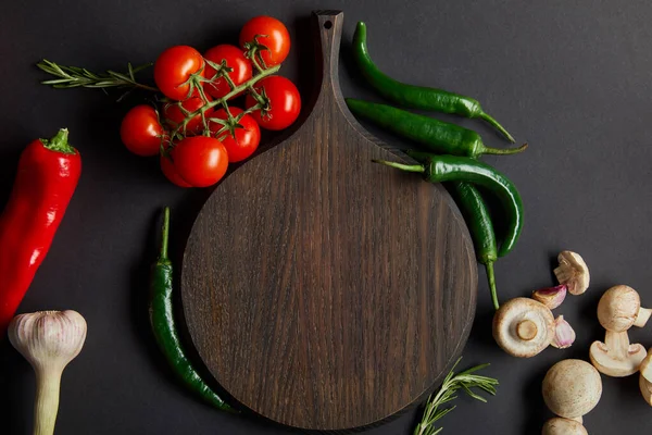 Top view of wooden cutting board near ripe cherry tomatoes, garlic, rosemary, green chili peppers and mushrooms on black — Stock Image Top View Wooden Cutting Board Ripe Cherry Tomatoes Garlic Rosemary Royalty Free Stock Images