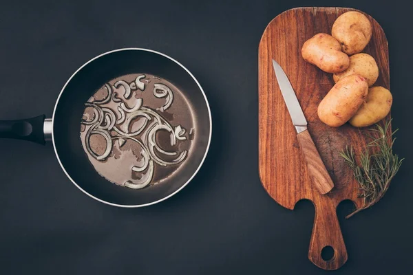 Top view of frying pan with onion and wooden board with potatoes on gray surface — Stock Photo