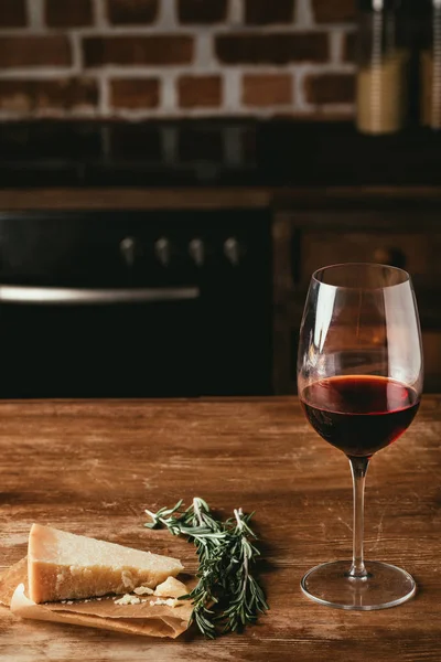 Copo de vinho tinto, queijo parmesão e alecrim fresco em mesa de madeira na cozinha — Fotografia de Stock