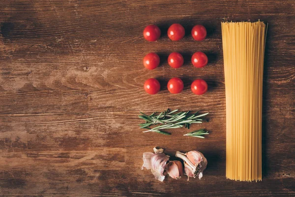 Plat posé avec des pâtes en rangée, tomates, romarin et ail sur une table en bois — Photo de stock