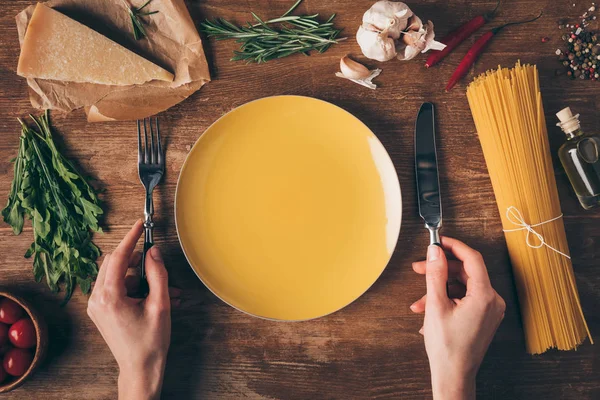 Vista recortada de las manos con cuchillo y tenedor en el plato con pasta de fila e ingredientes frescos alrededor de la mesa de madera - foto de stock