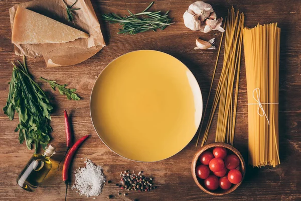 Vista superior de la pasta de fila, ingredientes frescos y plato en la mesa de madera - foto de stock